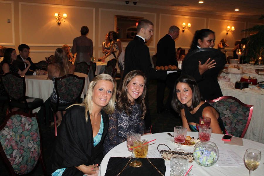 Three women smile at a table at a formal event, other guests in the background.