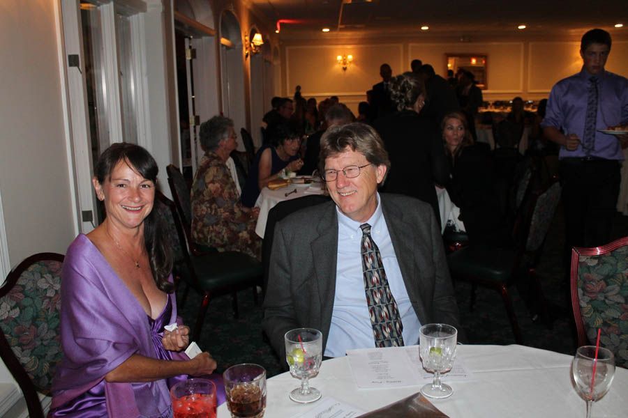 Woman in purple dress and man in suit at a table in a formal event; others in background.