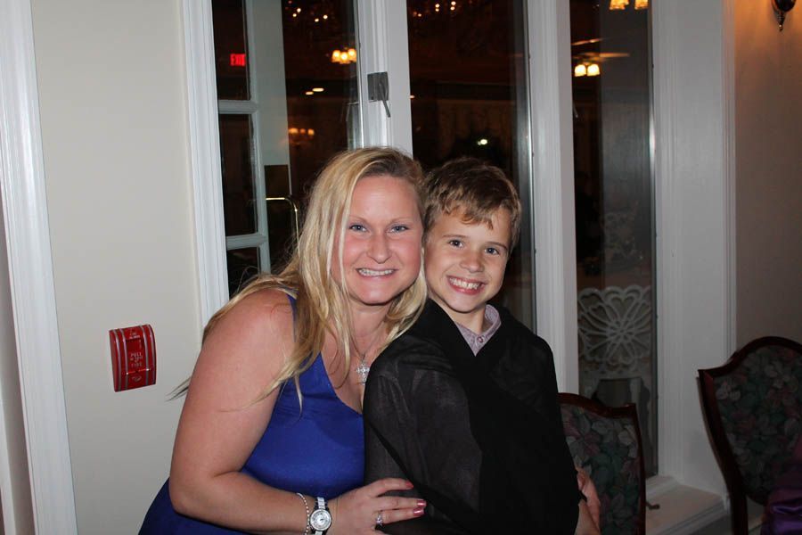 Woman in blue dress and boy in black outfit smile, posing by a window.