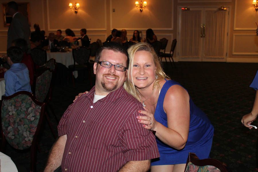 Man in maroon shirt and woman in blue dress smile, posing together in a banquet hall.