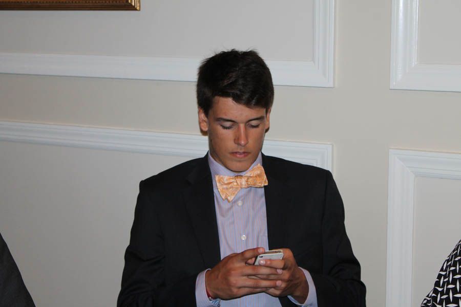 Man in suit with bow tie, looking at his phone. Set in room with white trim and wall.