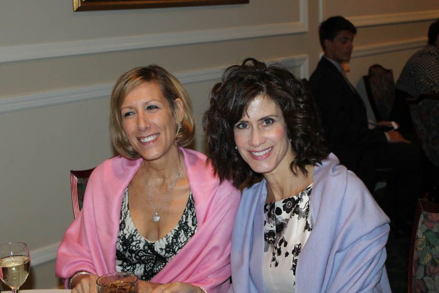 Two women smiling, seated. One in a pink shawl, the other in a purple shawl. Indoors, at a formal event.