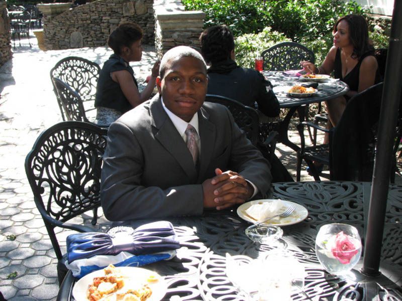 Man in a suit sits at a table outdoors, with other people dining in the background.