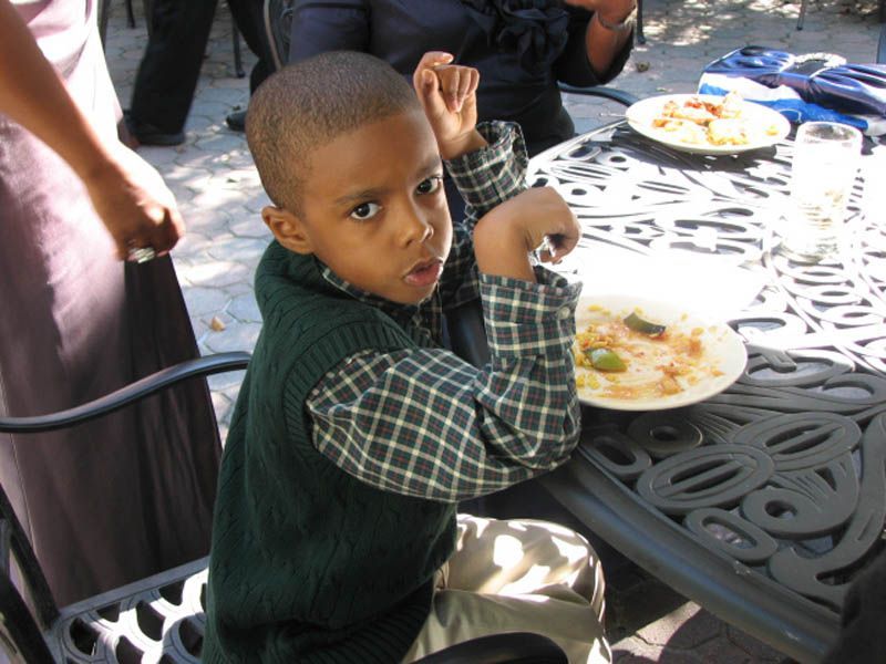 Young boy at a table, looking at the camera. Wearing a green sweater and plaid shirt. Outdoor setting, food on the table.