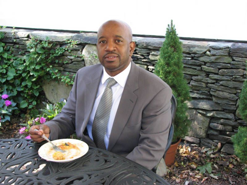 Man in suit eating at a table outdoors, small plate in front of him. Stone wall and plants in the background.