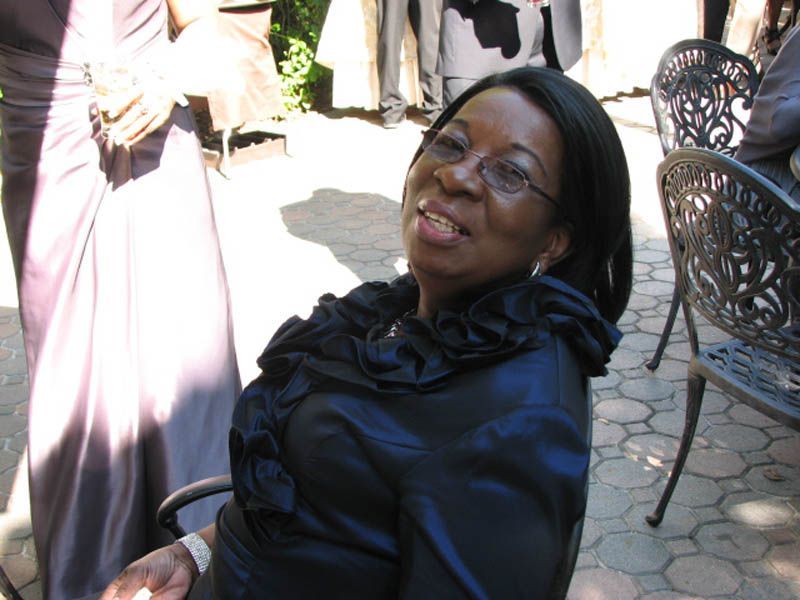 Woman smiling in a dark blue dress with a ruffled neckline, seated outside on a sunny day.
