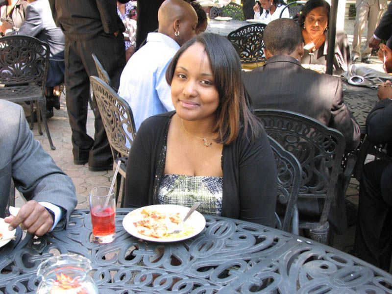 Woman at outdoor table, smiles, plate of food, drink, ornate metal chairs. Other people in the background.
