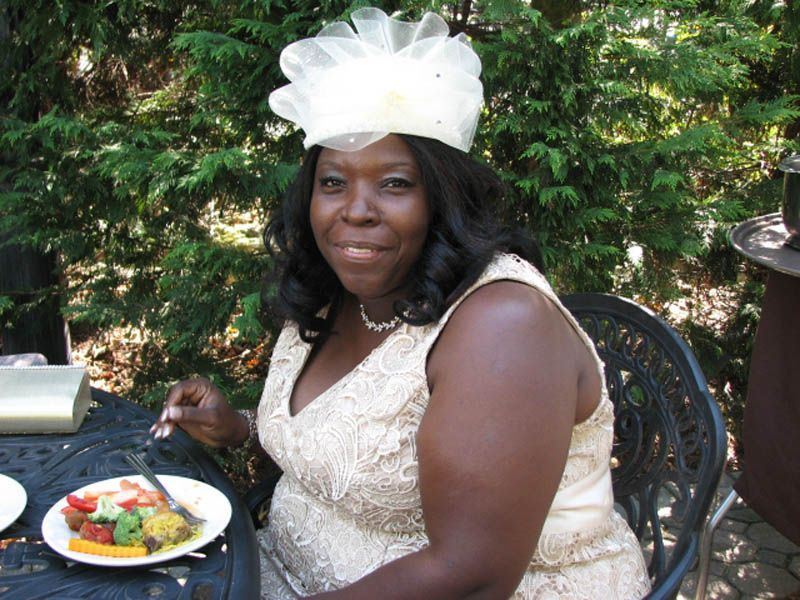 Woman in cream-colored dress and hat smiles, seated outdoors at a table with food.