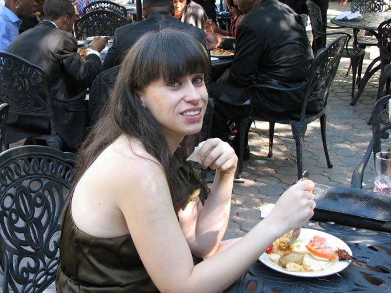 Woman eating at outdoor restaurant, holding food and a napkin, smiling. Others seated at tables.