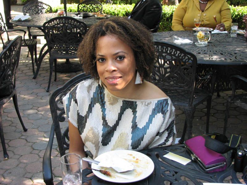 Woman seated at outdoor table, smiling. Wearing patterned top, eating from plate. Black wrought iron furniture.