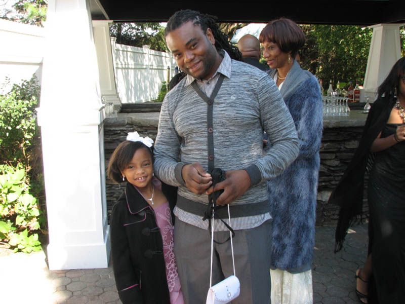 Man and girl pose outdoors, others in background. Man wears gray sweater, girl in coat, bow.