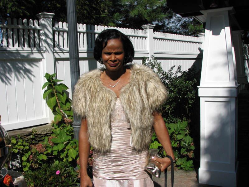 Woman in a cream-colored dress and fur shawl stands outdoors in front of a white picket fence and greenery.