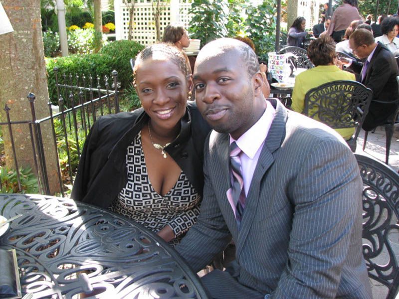 Couple smiles at outdoor cafe table; woman in patterned top, man in suit.