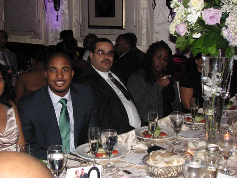 People seated at a formal table at an event. A man in a suit smiles. Flowers and glassware are on the table.