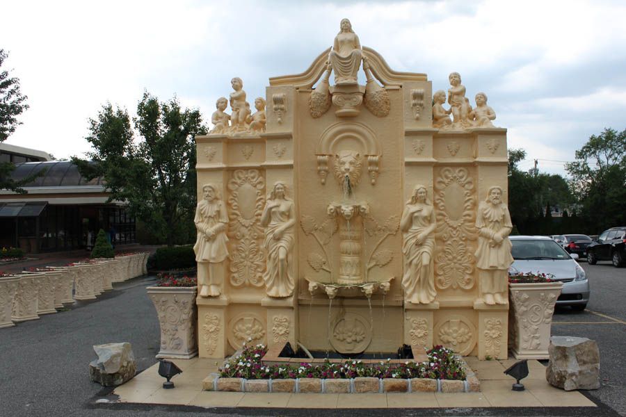Ornate beige fountain with sculptures, flower beds, and cars in a parking lot.