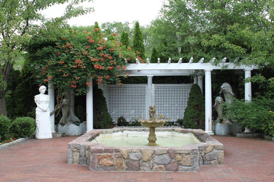 Stone fountain in a brick-paved garden with a trellis, statues, and flowering vines.