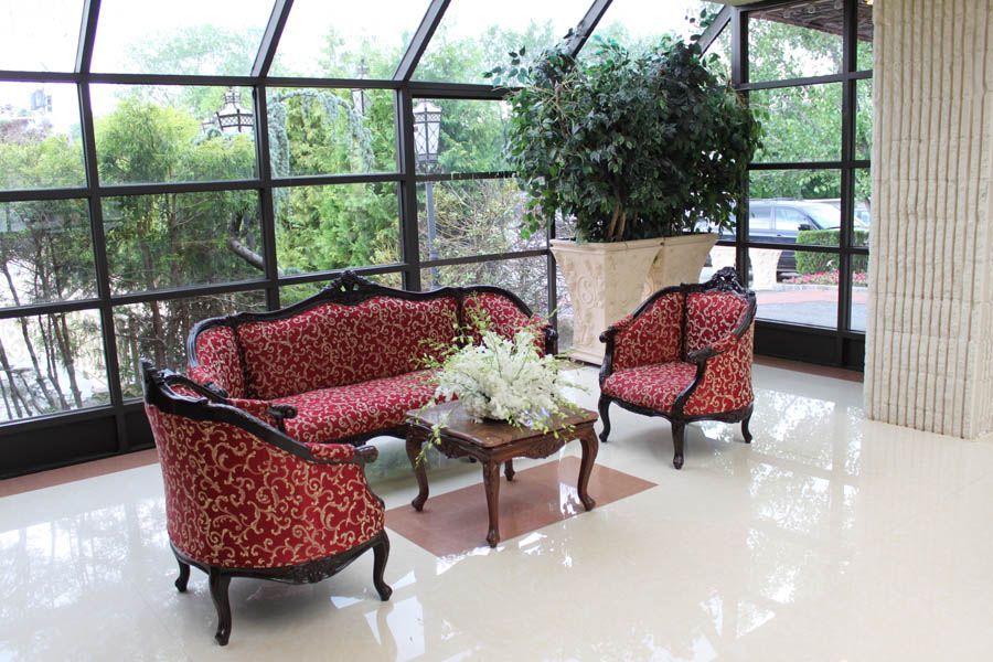 A waiting area with red patterned furniture, a small table, and a large plant. Windows overlook greenery and a street.