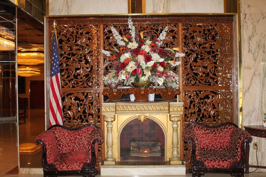 Ornate hotel lobby with a fireplace, floral arrangement, and two red patterned armchairs; US flag.