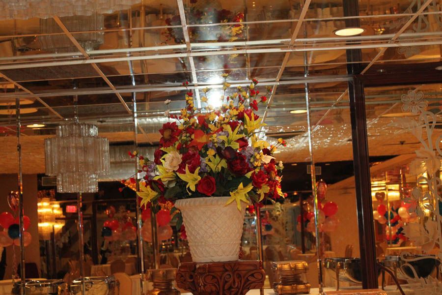 Floral arrangement in a white vase on ornate stand, in a mirrored room with chandeliers and party decor.