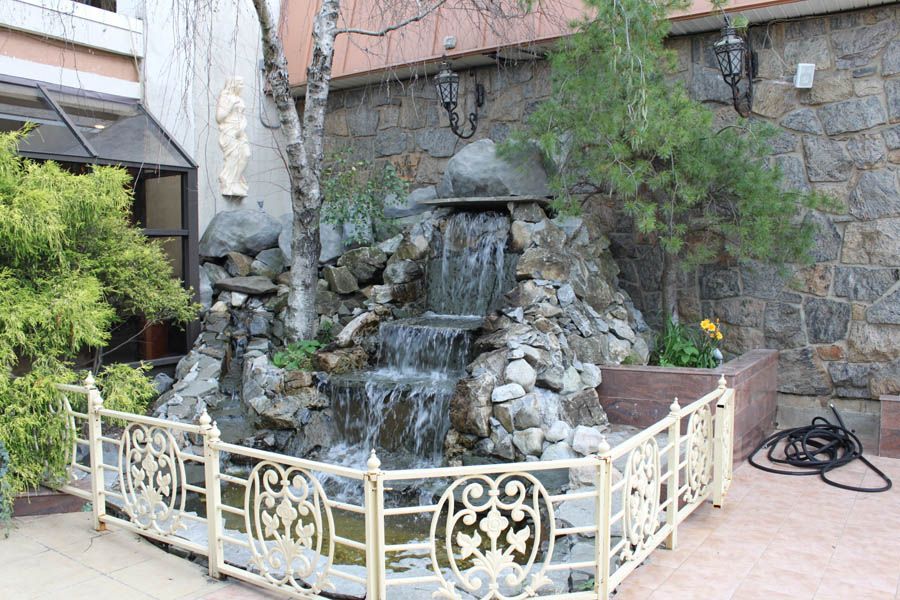 Waterfall feature in a stone-walled outdoor area, with ornamental white railing in the foreground and a hose on the right.