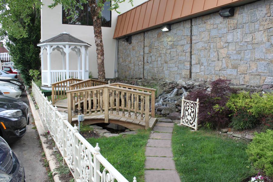 Wooden bridges over a small stream, leading to a gazebo, next to a building with a stone wall and landscaping.