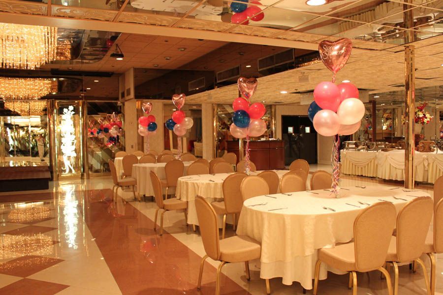 Banquet hall with round tables set for a party, decorated with balloon bouquets in pink, white, and blue.