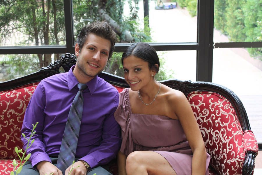 Couple sitting on a red patterned couch, smiling. Man in purple shirt/tie, woman in lavender dress.