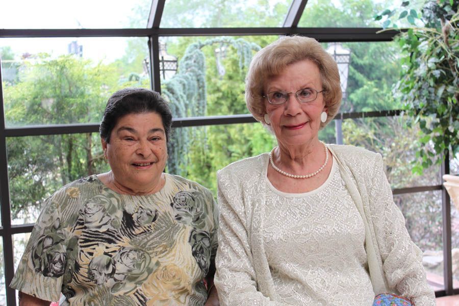 Two women smiling in front of a window. One in a patterned top, the other in a sparkly white outfit with a necklace.