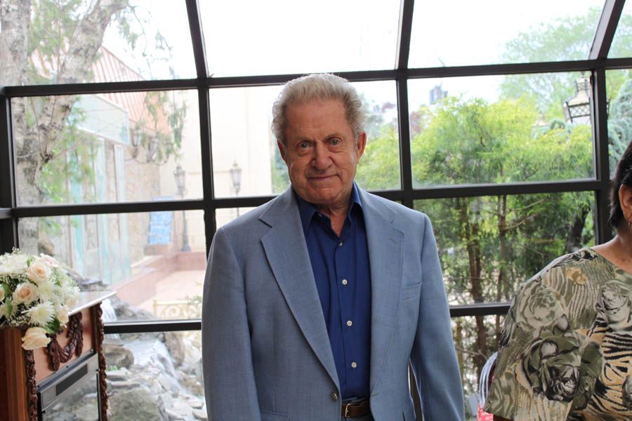 Man in a light blue suit and blue shirt stands by a window in a bright setting.