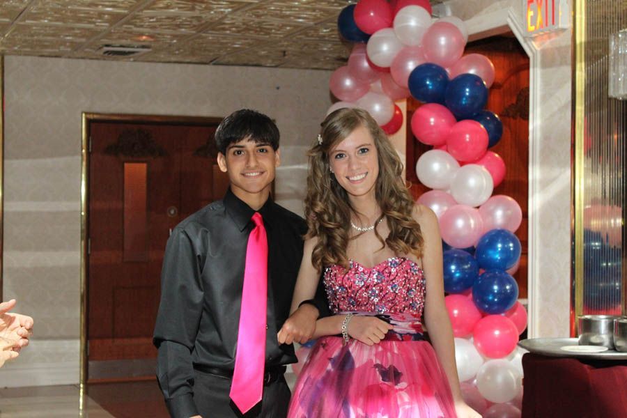 Couple at a formal event; boy in grey shirt, pink tie, girl in pink sequin dress; pink, blue, white balloon arch.