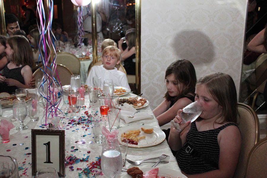 Children at a decorated banquet table with food and drinks. One girl drinks water. Confetti and decorations are present.