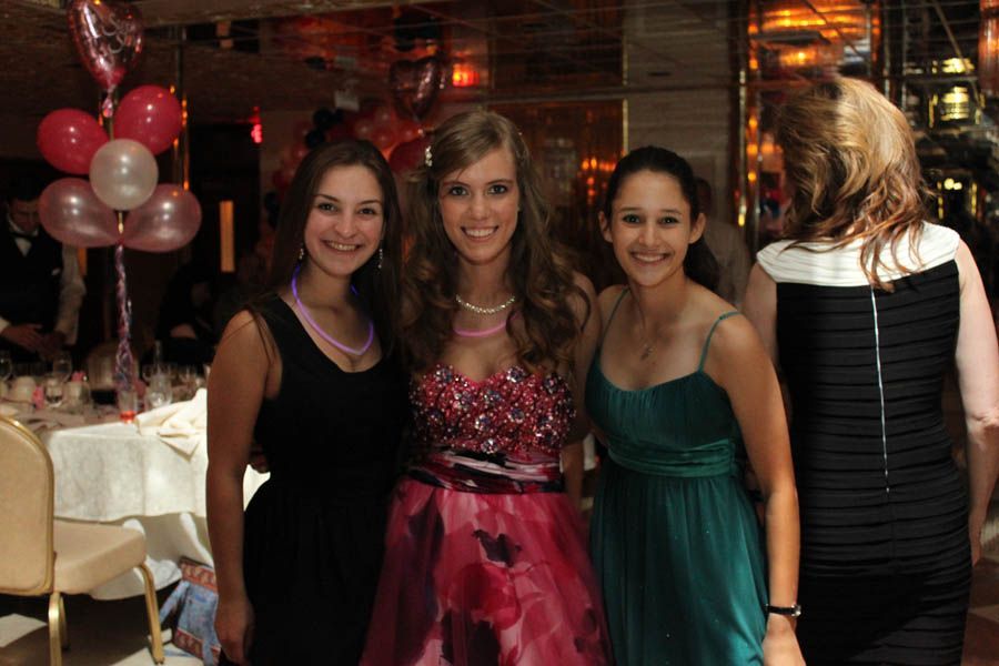 Three young women in formal dresses pose at a celebration, with balloons and tables in the background.