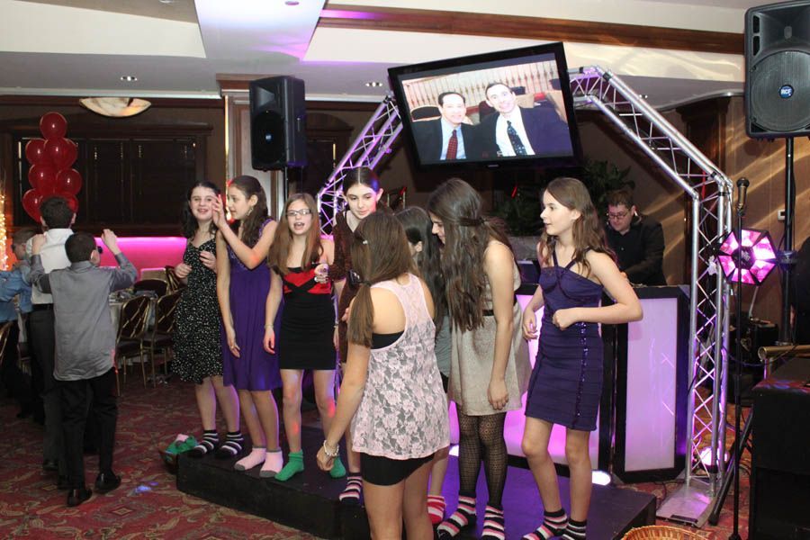Teenagers singing at a party, on a stage with a screen showing a video. DJ booth and speakers on the side.