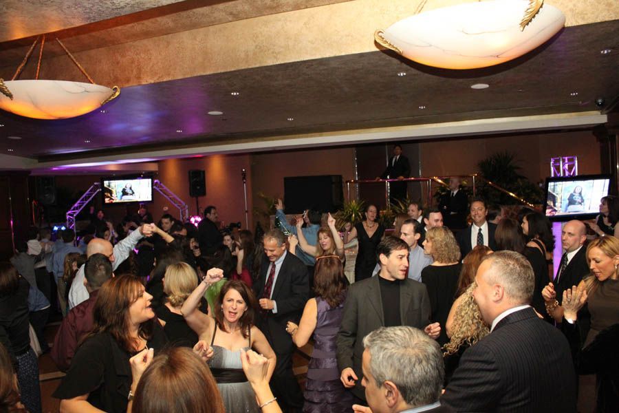 People dancing in a well-lit ballroom with wall-mounted TVs. Some wear formal attire.
