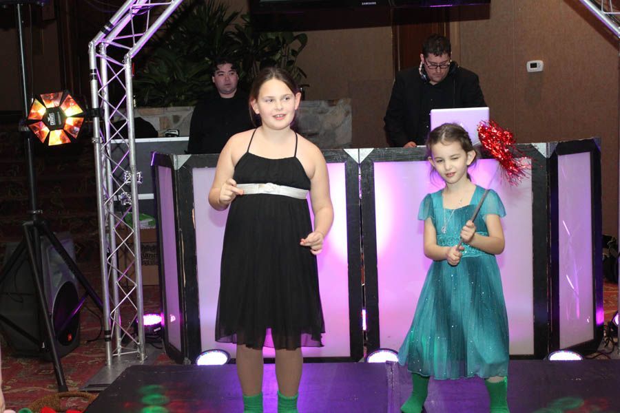 Two girls dance on stage in front of a DJ setup, a red bow in one's hair.