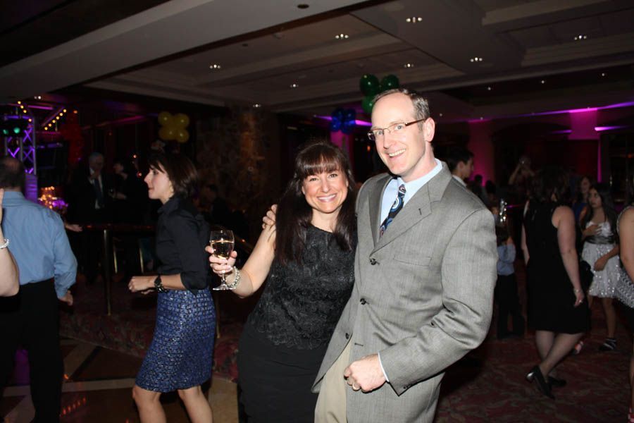 Couple dancing at a party; woman holds a drink, man smiles. People and decorations in the background.