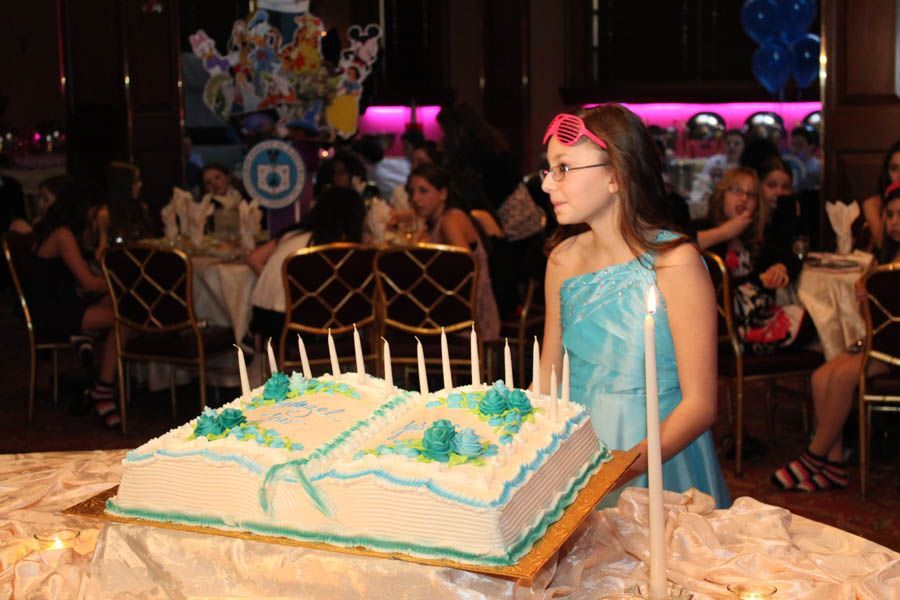 Girl in a blue dress with pink glasses by a cake shaped like an open book, with candles, in a party hall.