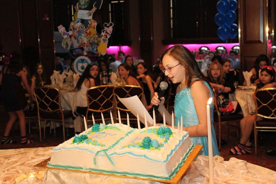 Girl in blue dress speaking at a party, holding paper near a cake shaped like a book with candles.