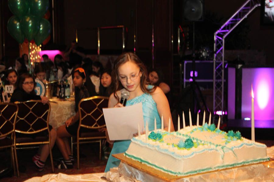Girl in blue dress speaking at a party, cake with candles in foreground, guests seated at tables.