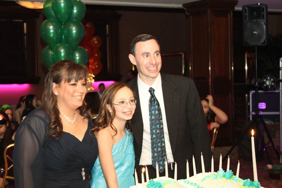 Family at a party with a cake. Woman in navy dress, girl in blue, man in suit. Balloons in background.