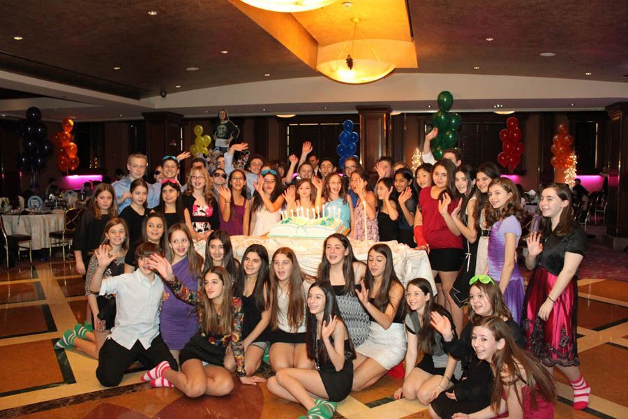 Large group of young people celebrating around a cake in a ballroom, with balloons and formal attire.