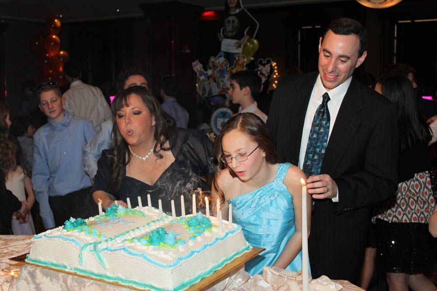 Family blowing out candles on a cake at a party, illuminated by warm lights.