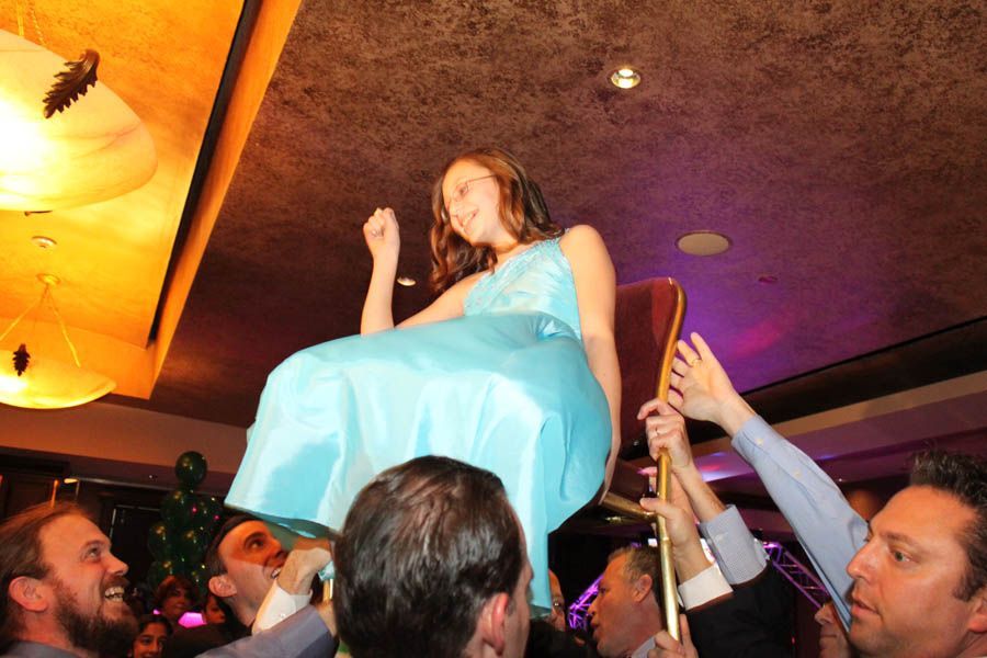 Person in blue dress is lifted on a chair by a group of people, celebrating. Party setting with lights.
