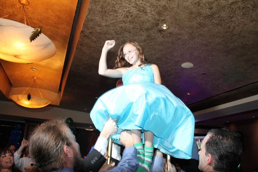 Girl in blue dress being lifted on chairs at a party, smiling and raising her arm.