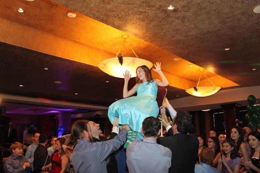 Girl in turquoise dress lifted on chairs at party. People cheering under warm lighting.