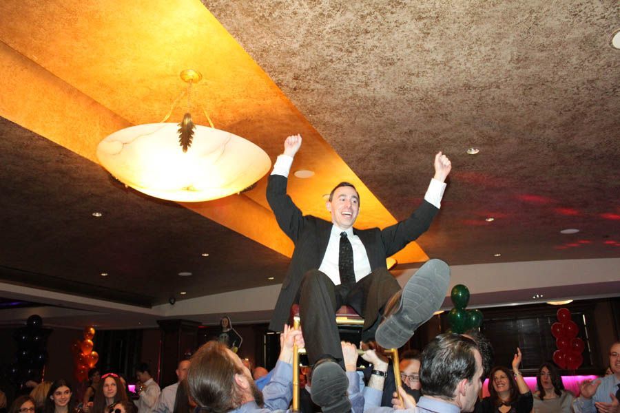 Man in suit raised in the air by a crowd at a party, arms raised in celebration, under a large light fixture.