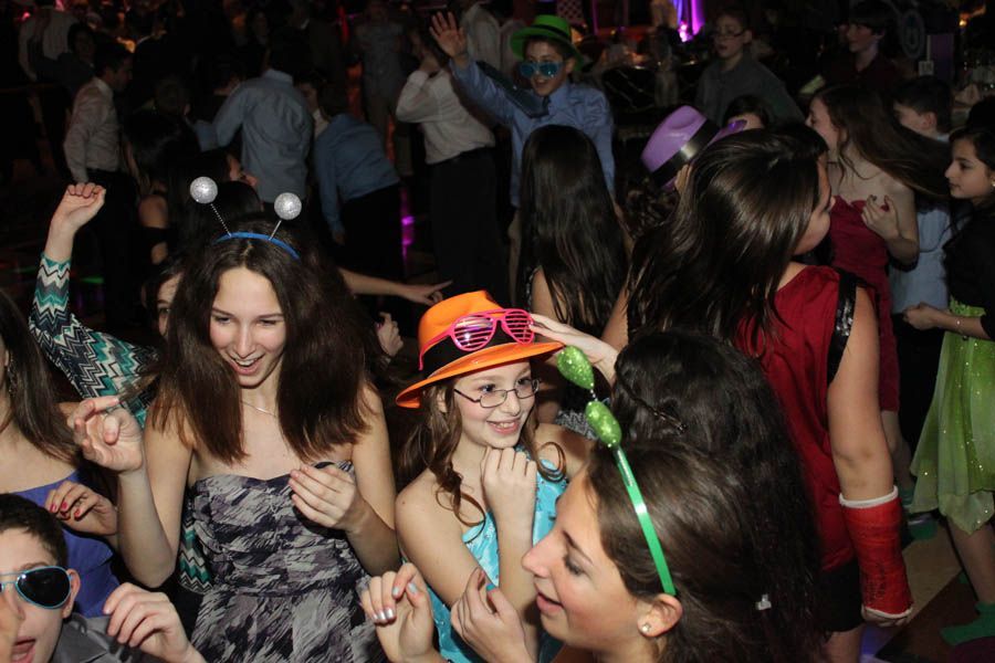 People dancing at a party, wearing hats and accessories. Dimly lit with flashing lights.
