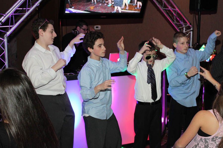 Four boys dancing at a party with a DJ booth in the background, lit with pink and purple.