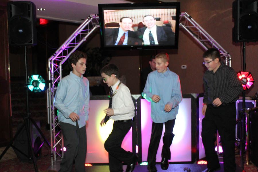 Four boys dancing in front of a DJ booth with lights and a screen showing two men.