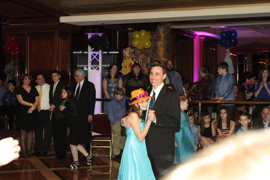 Man and girl in teal dress dance at a party; people watch, some with balloons and festive lighting.